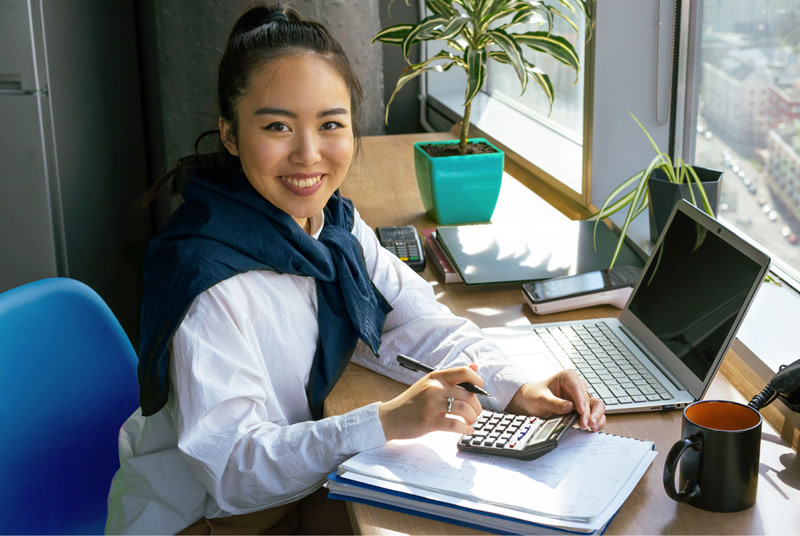 A bookkeeper smiling at her desk with a calculator, ledger and laptop